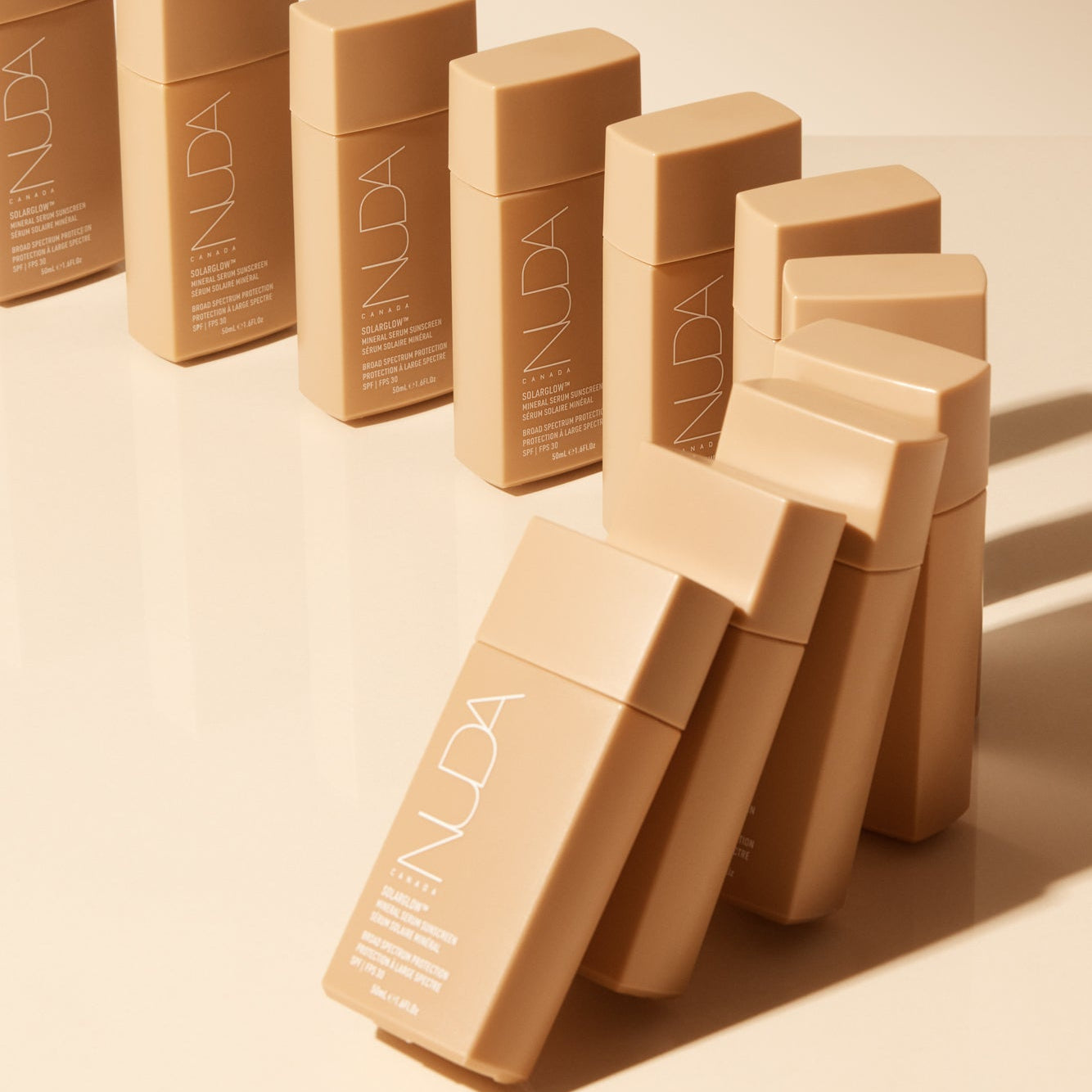 A group of NUDA liquid foundation bottles in a warm beige shade, arranged diagonally on a matching beige studio background.