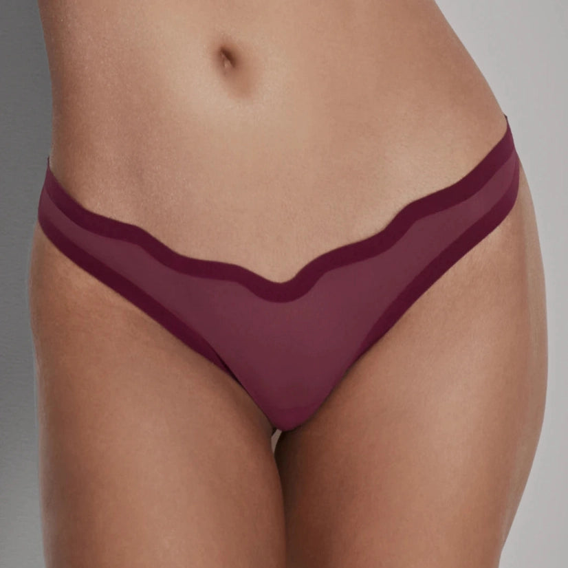 A close-up of a model's lower torso wearing a sheer burgundy mesh thong with solid burgundy trim and a scalloped wavy neckline.