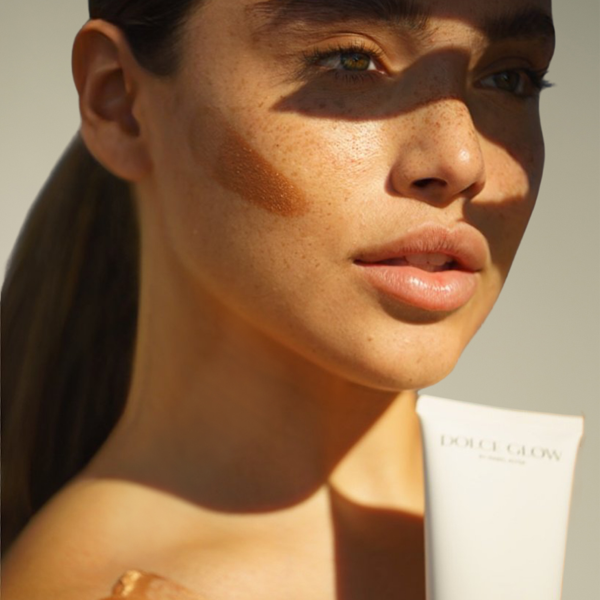 Close-up of a woman's face with a tube of Dolce Glow product and a swatch of the product on her cheek