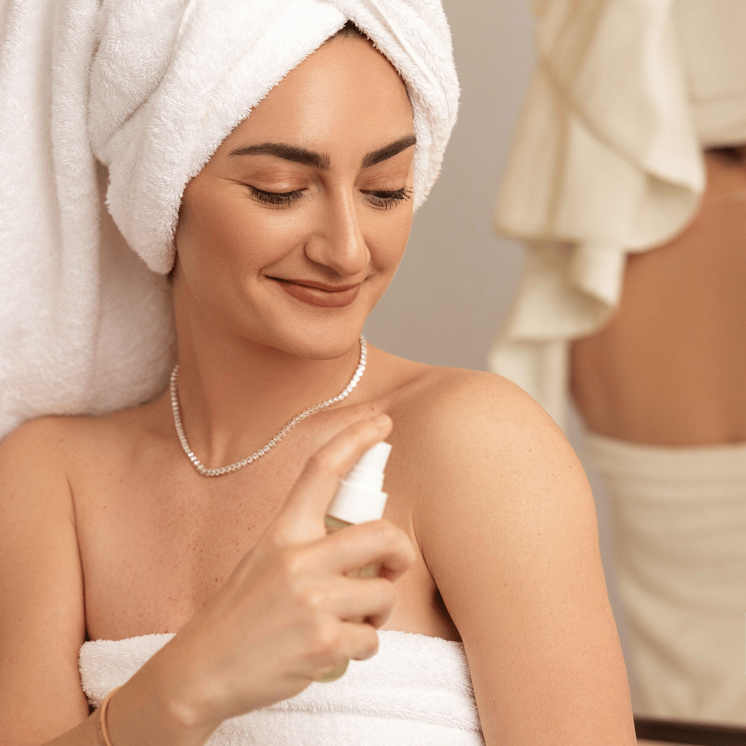 Woman applying spray to her arm while wearing a towel on her head, in a bathroom setting.