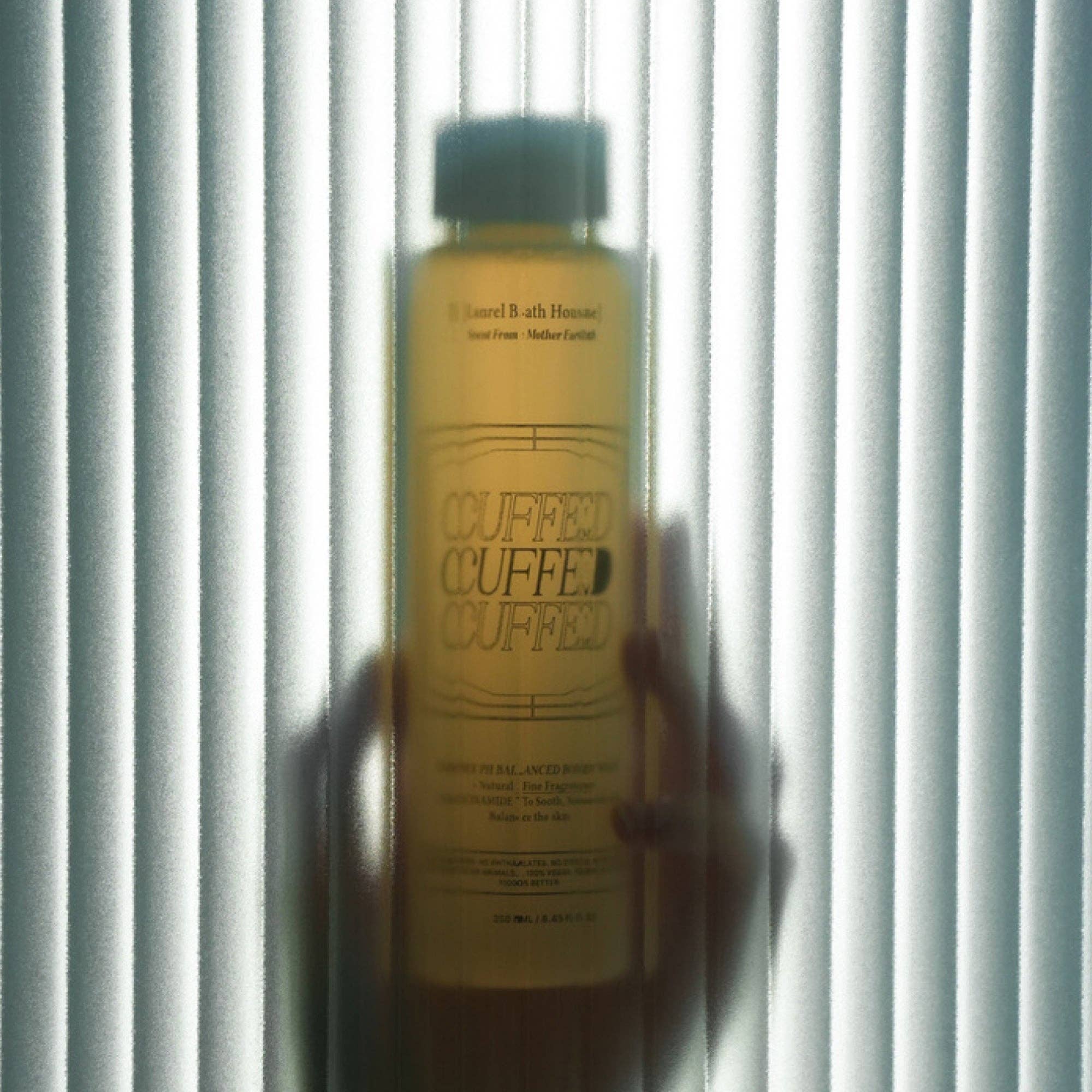 Bottle of 'Cuffed' body wash held against a striped background