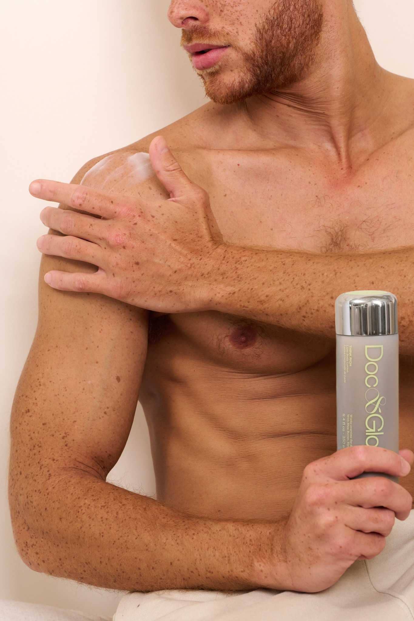Man applying Think Rich Luxe Body Lotion to his shoulder holding a bottle on a white background. 