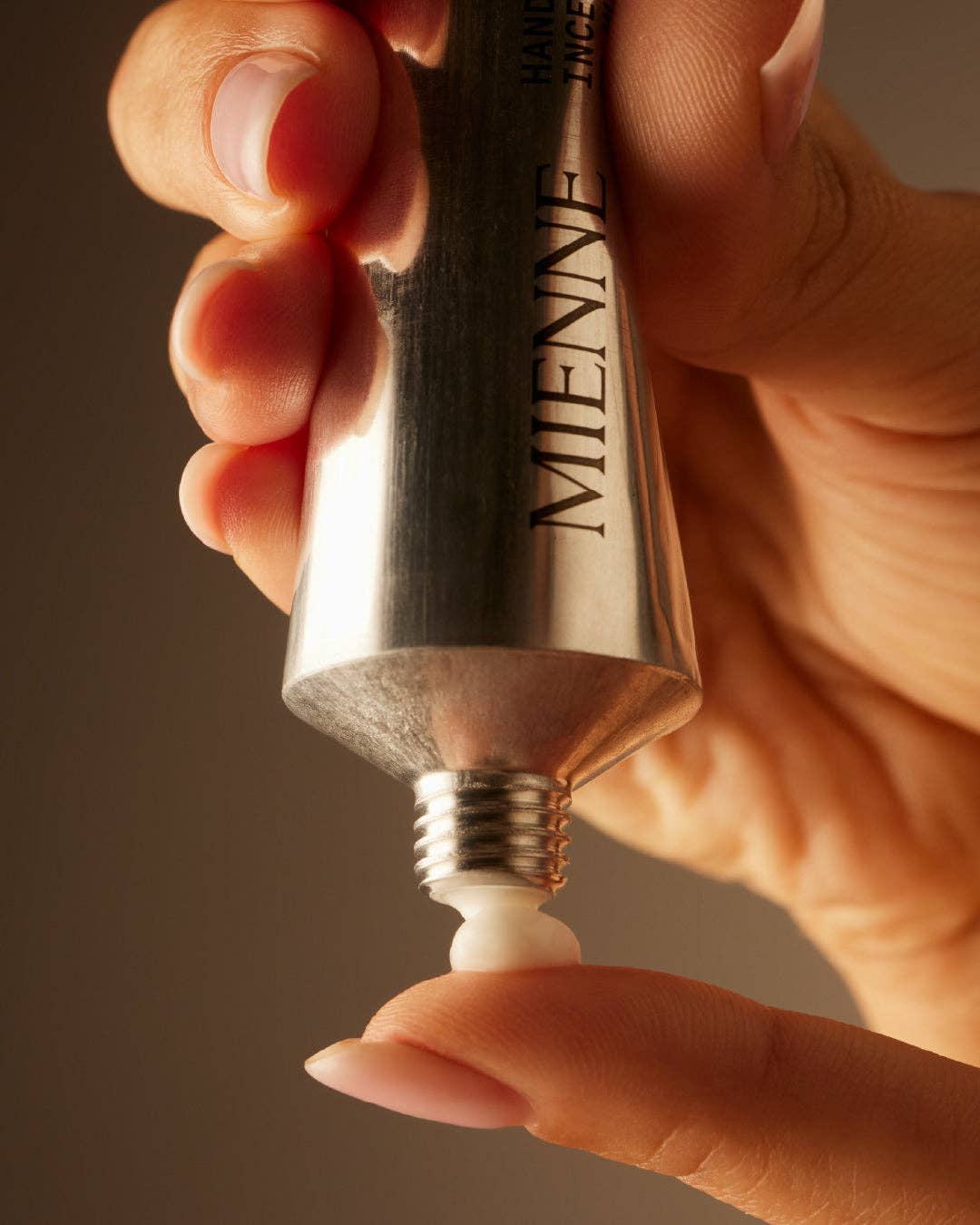 Hand holding a silver Miwene lip balm against a brown background