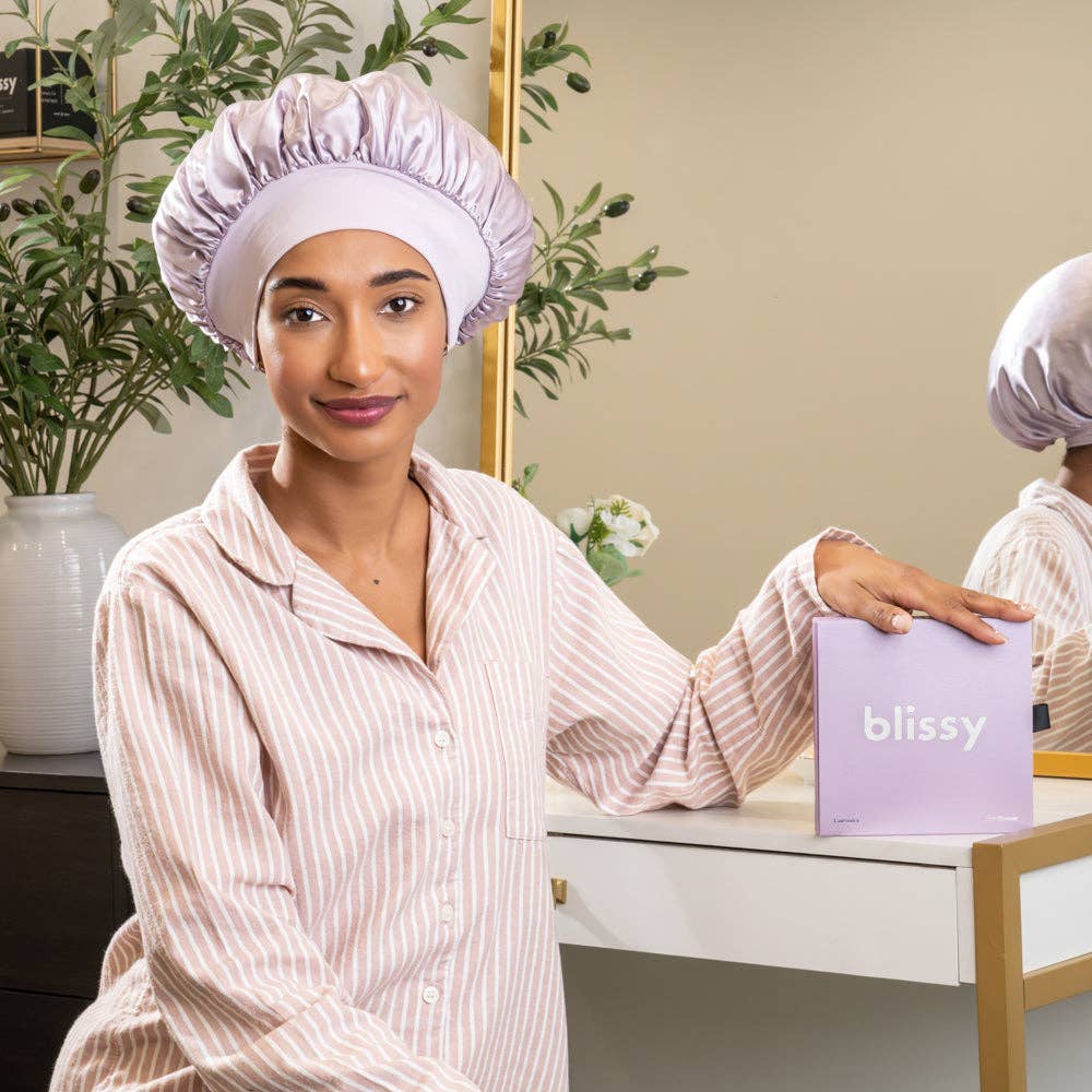 Woman wearing a satin hair cap and holding a 'blissy' product box in front of a mirror.