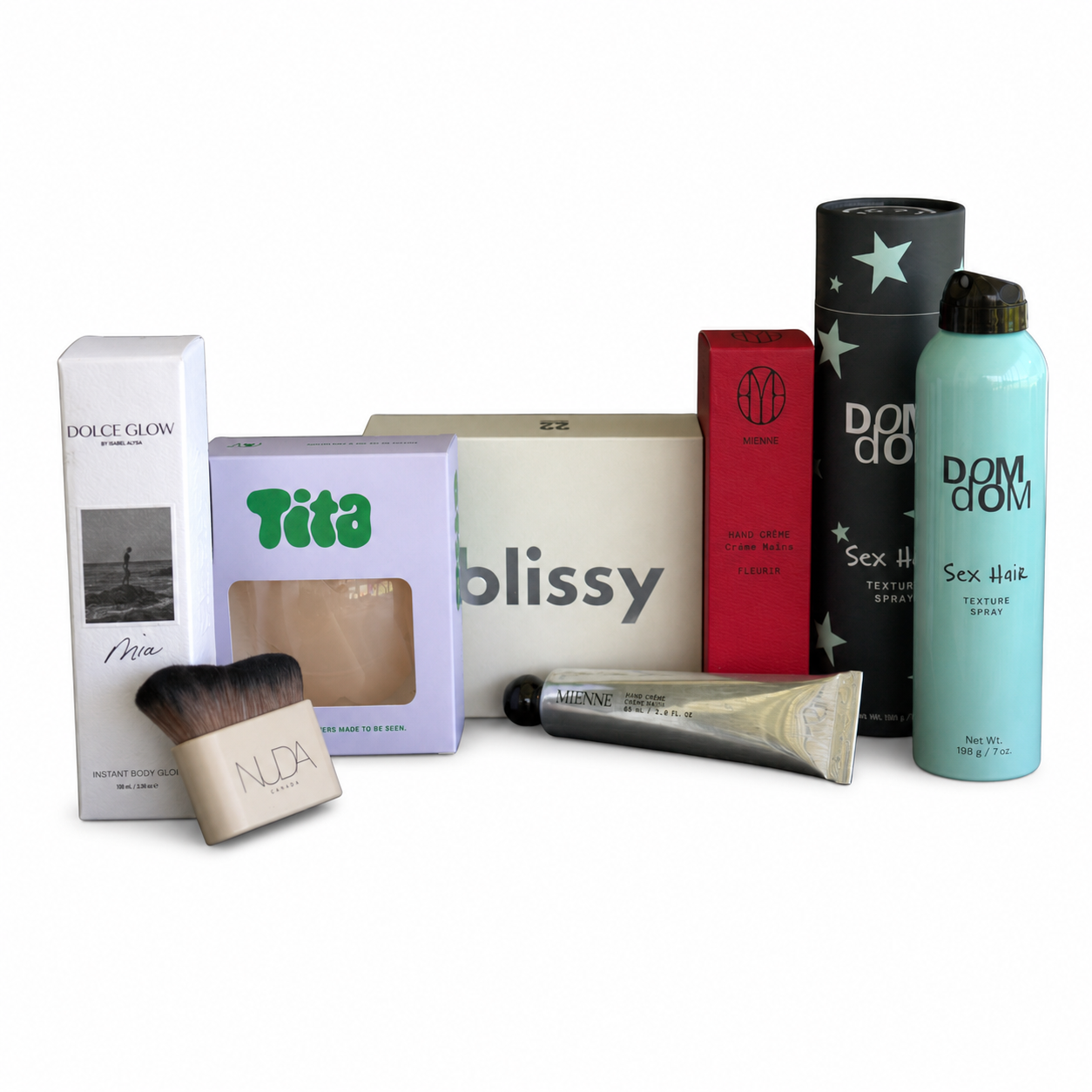 A collection of beauty and haircare products arranged together on a white background, including a white Dolce Glow perfume box, a lavender Tita hair color box, a cream-colored blissy box, a red box, a black star-patterned tube, a light blue Dom Glow Sex Hair spray bottle, a silver tube, and a beige makeup brush.