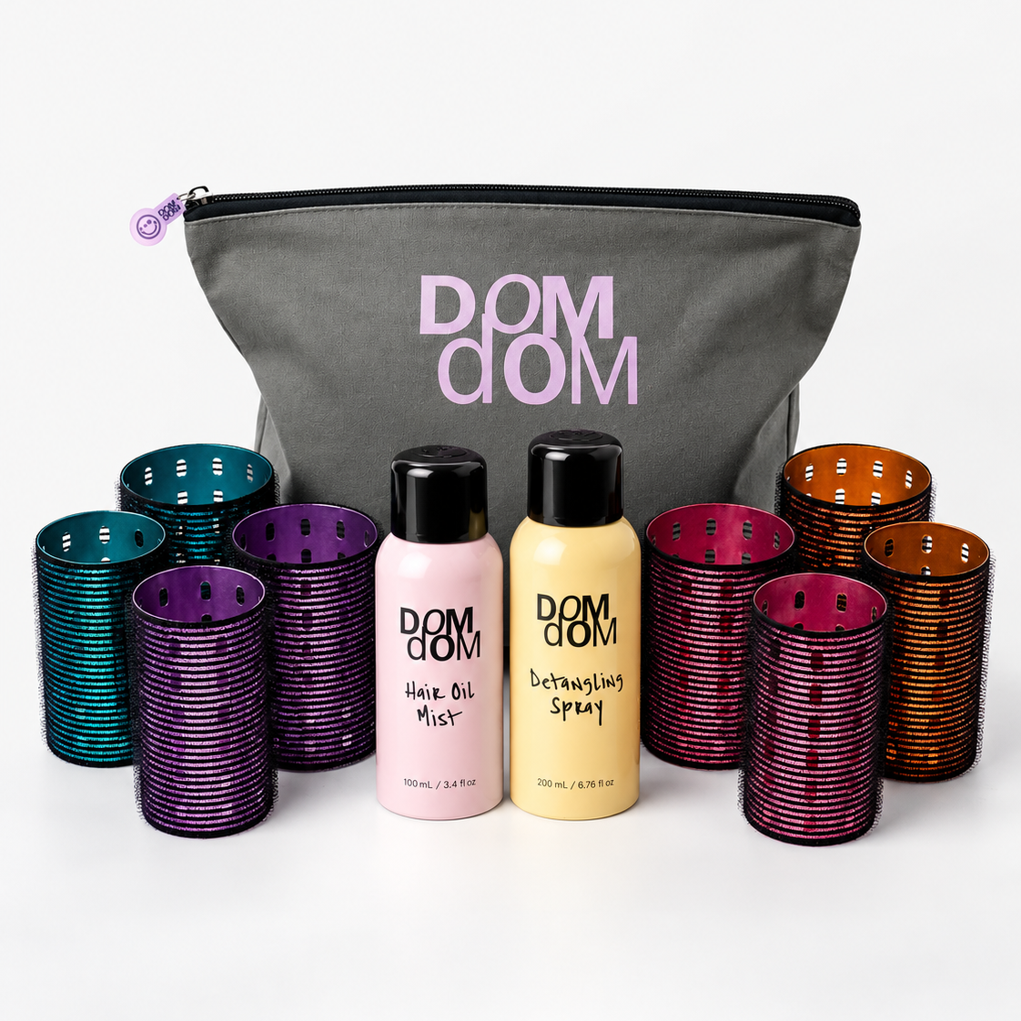 Dom Dom hair care set featuring a gray zippered pouch with pink lettering, two bottles of hair product (Hair Oil Mist in pink and Detangling Spray in yellow), and multiple stacks of colorful silicone hair rollers in purple, teal, magenta, and brown, arranged on a white background.