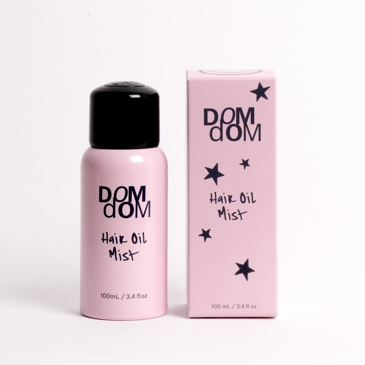A pink bottle of DOM DOM Hair Oil Mist with a black cap stands next to its matching pink packaging box on a clean white background.