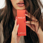 Person holding a red 'Mane Ivy' hair serum box with long dark hair.