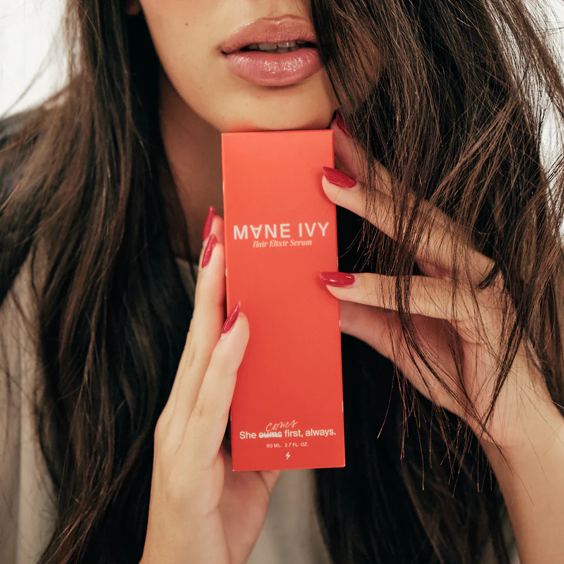 Person holding a red 'Mane Ivy' hair serum box with long dark hair.