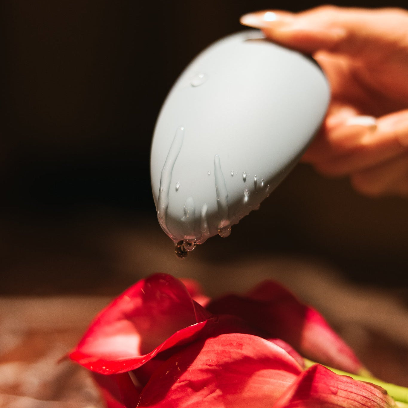 hand holding a blue massager with liquid dripping off onto red flowers with a dark background. 