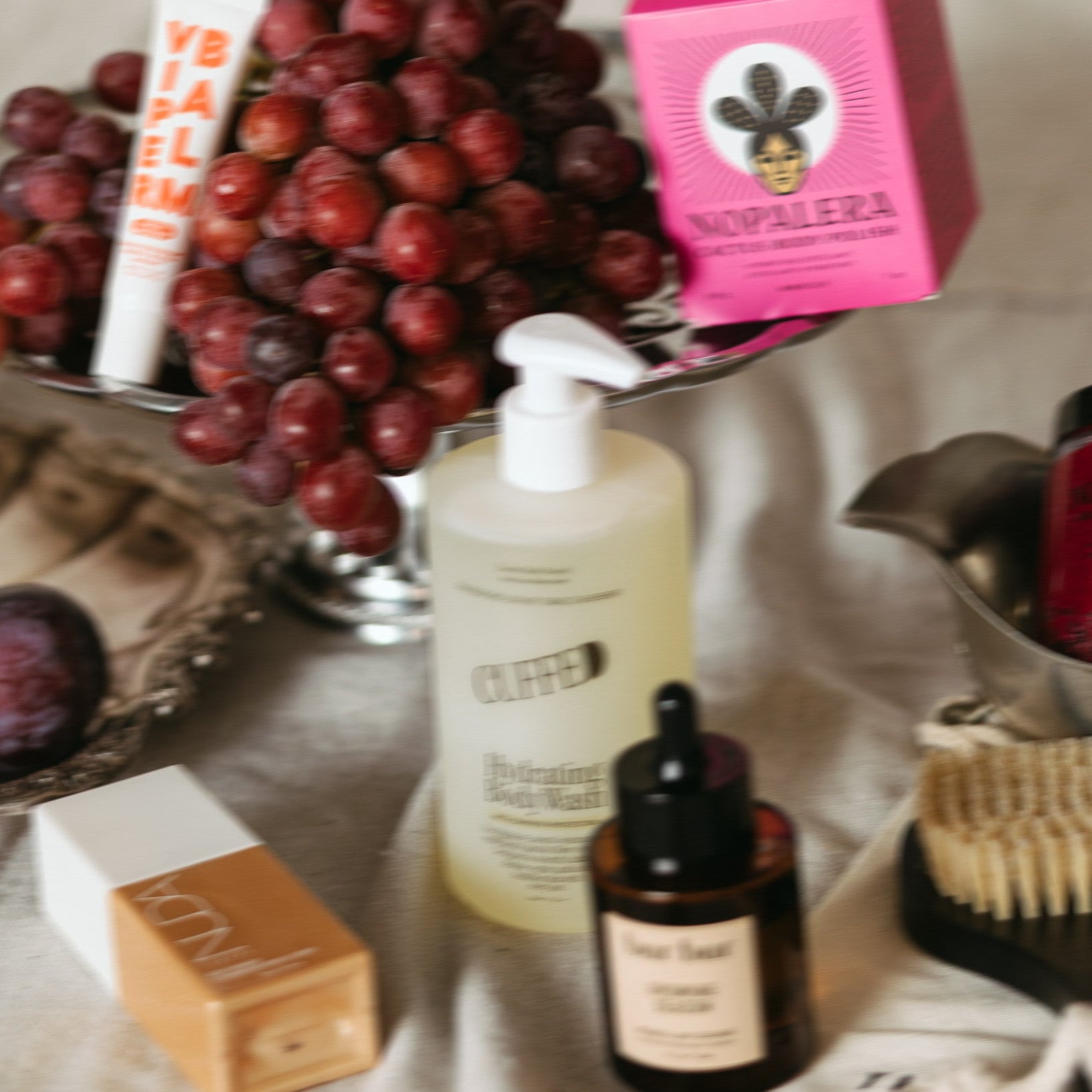 Cosmetic products including a bottle, dropper, and soap on a textured surface with grapes in the background.