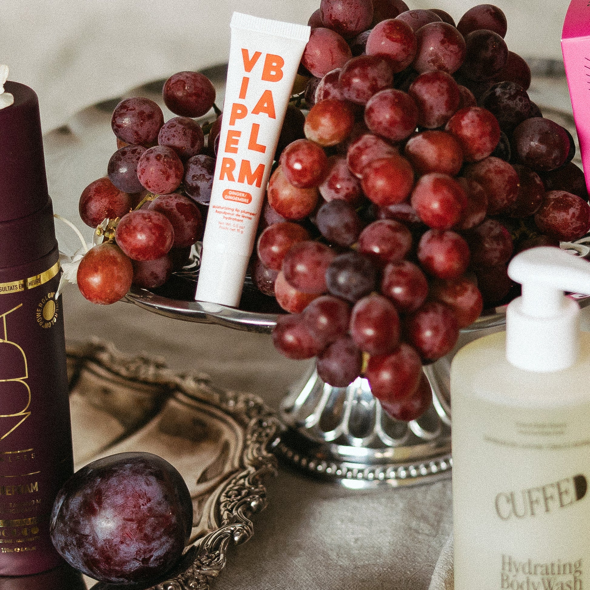 Viper balm tube,  bottle of body wash, and plum on a decorative stand with grapes.