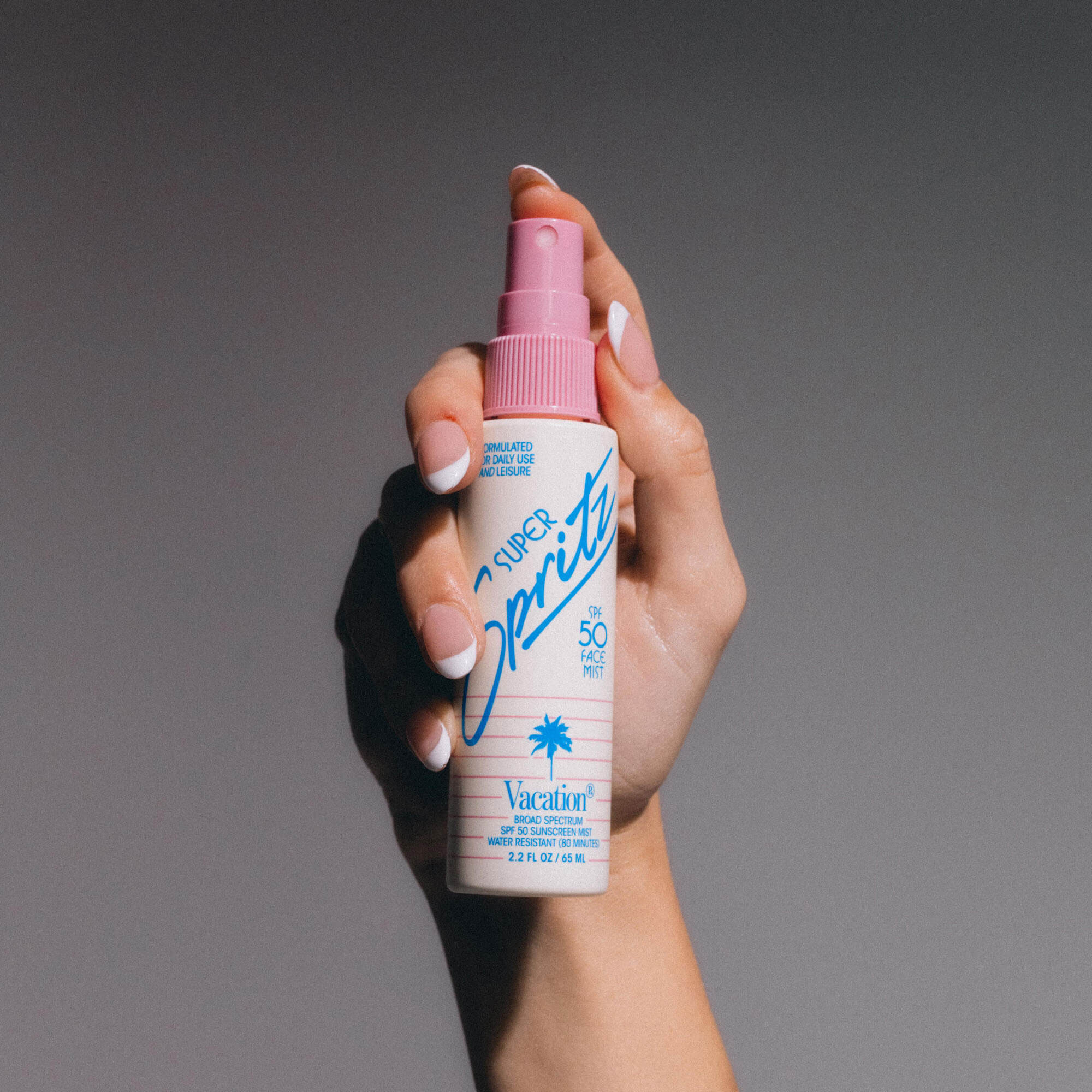Hand holding a Super Spritz sunscreen spray bottle with pink nozzle against a gray background
