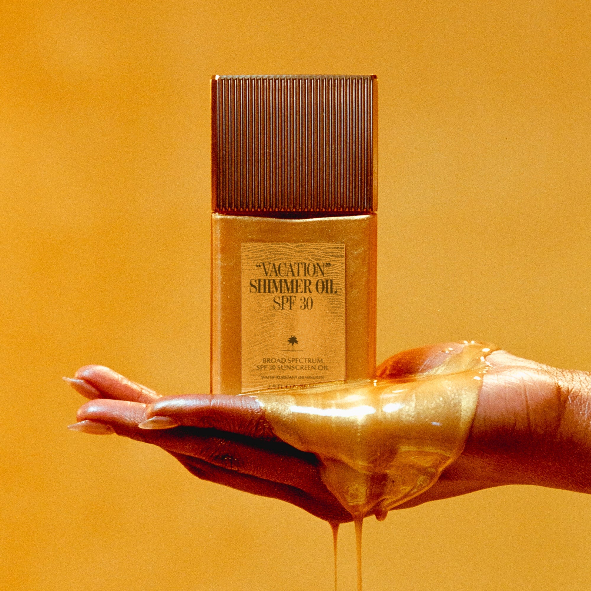Gold bottle of 'Vacation Shimmer Oil SPF 30' held by a hand with a yellowish-orange background
