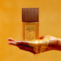 Gold bottle of 'Vacation Shimmer Oil SPF 30' held by a hand with a yellowish-orange background